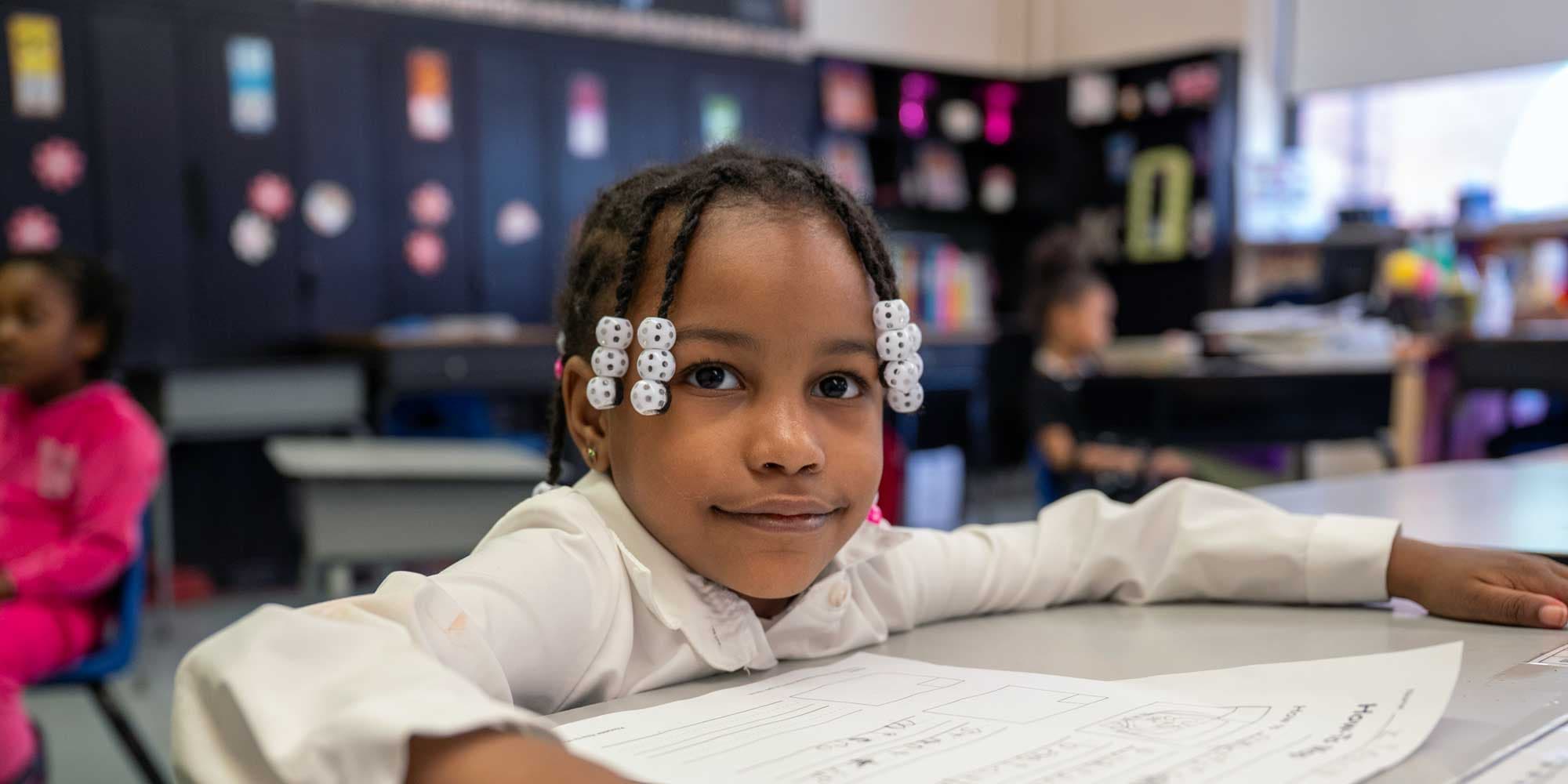 Young student with braided hair and beads sitting at a classroom desk.