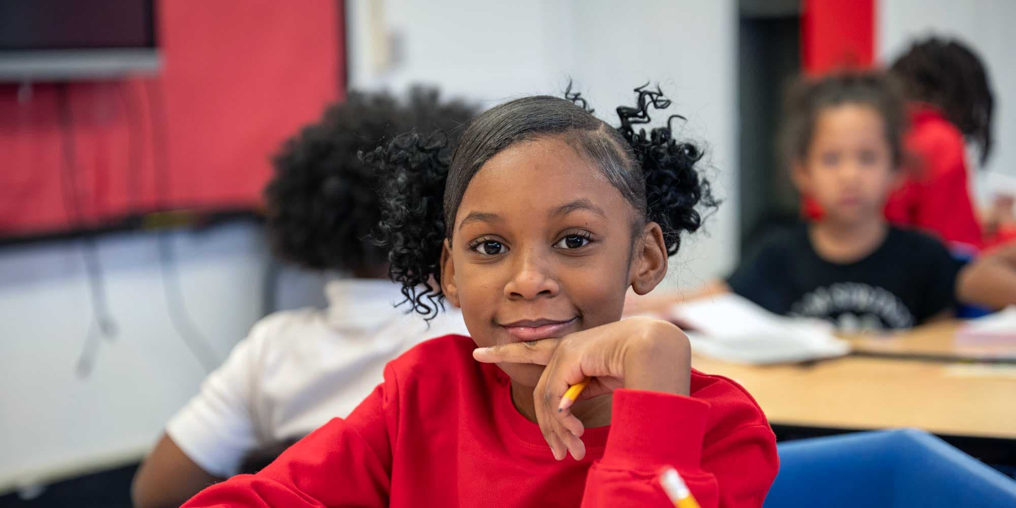 Smiling student in a red shirt holding a pencil at a desk.