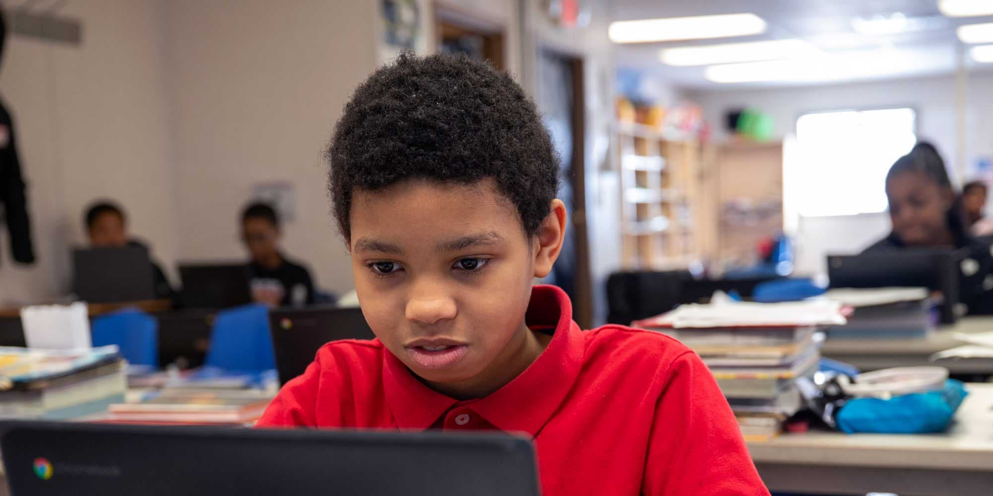 Student in a red shirt focused on a laptop in a classroom.