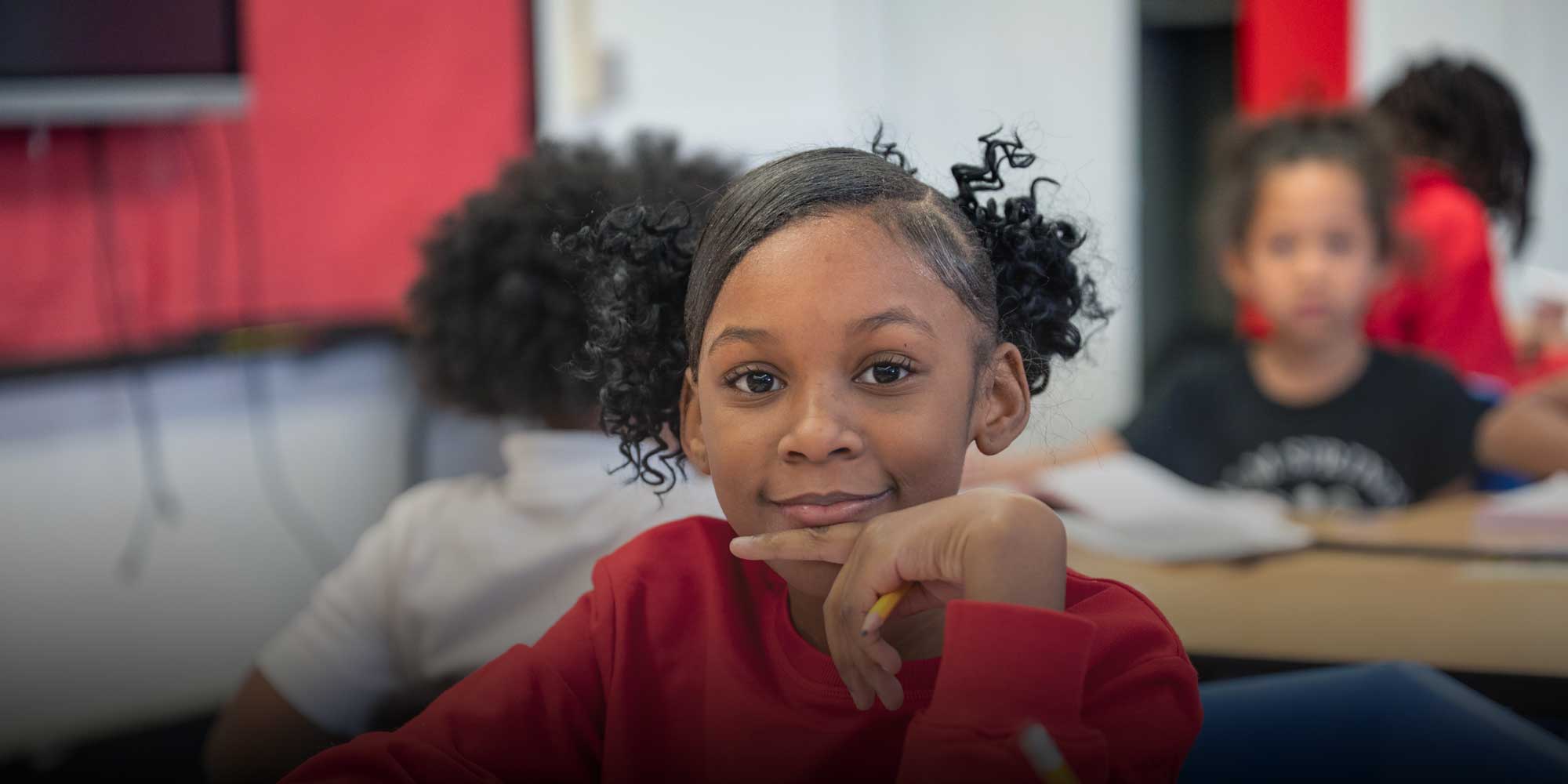 Smiling East Academy student in a red shirt resting their chin on a hand at a desk.